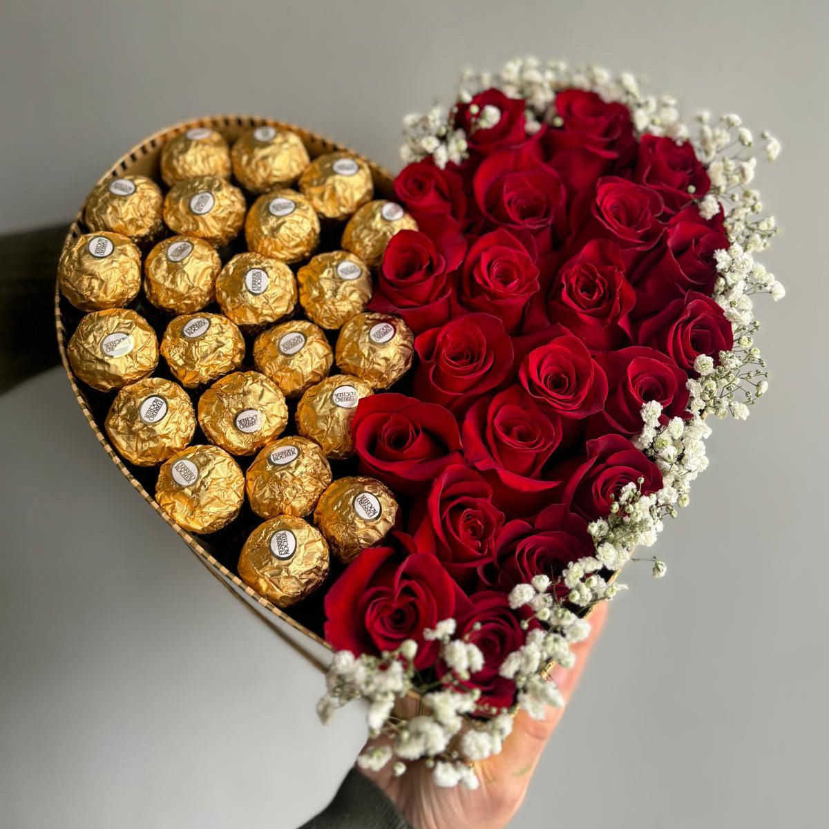 Heart-shaped arrangement of gold Ferrero Rocher chocolates and red roses on a gray background