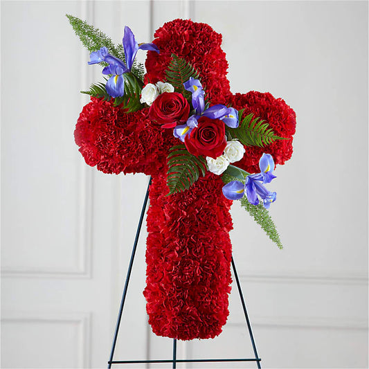 Cross-shaped floral arrangement made with red carnations, accented by red roses, white spray roses, blue irises, and lush greenery, displayed on a black stand. A vibrant and meaningful tribute symbolizing love, faith, and remembrance for a funeral or memorial service