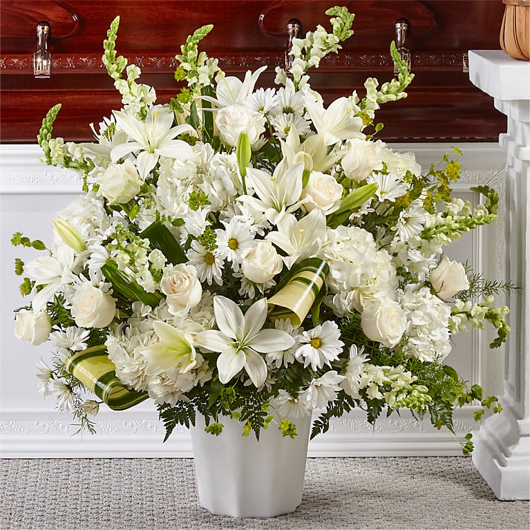 Bouquet of white flowers in a white vase in front of a wooden casket.