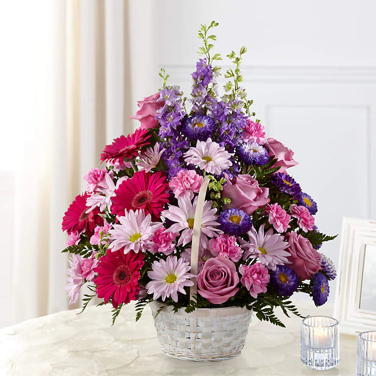 Bouquet of pink, purple, and white flowers in a white basket on a light surface with a white curtain background.
