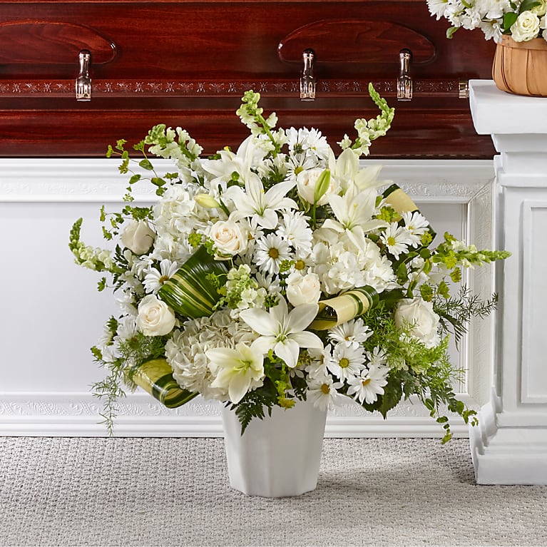 White floral arrangement in a vase in front of a white casket with wooden trim.