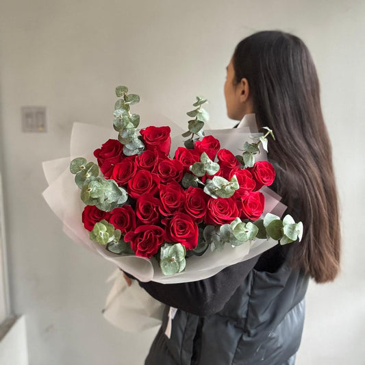 Person holding a bouquet of red roses with greenery against a plain background