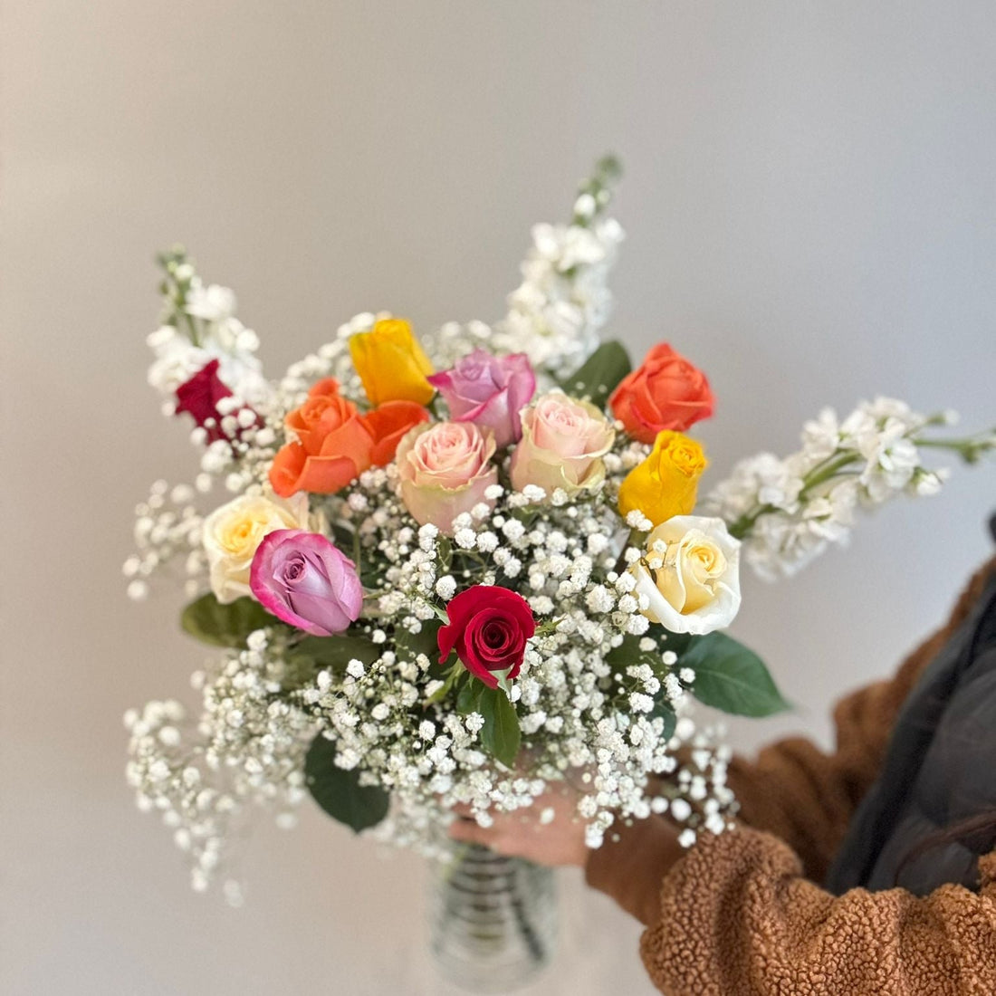 Bouquet of colorful flowers held by a person wearing a brown jacket against a neutral background