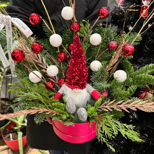 "A compact holiday flower box featuring vibrant red poinsettias, white chrysanthemums, and lush evergreen sprigs, accented with pinecones and a red ribbon. The arrangement is displayed in a rustic wooden box, perfect for festive decor or gift-giving."