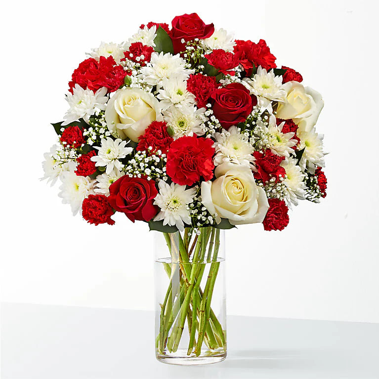 A vibrant winter fiesta bouquet in a clear glass vase, featuring a mix of red roses, red carnations, white roses, white chrysanthemums, and baby's breath. Perfect for festive holiday decor.

