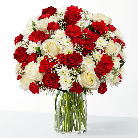 A vibrant winter fiesta bouquet in a clear glass vase, featuring a mix of red roses, red carnations, white roses, white chrysanthemums, and baby's breath. Perfect for festive holiday decor.