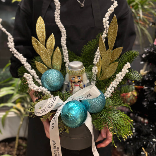 A festive Christmas flower box filled with red poinsettias, white chrysanthemums, and greenery, decorated with pinecones and red ribbon. The arrangement is placed in a wooden box with a rustic finish, evoking a cozy holiday atmosphere