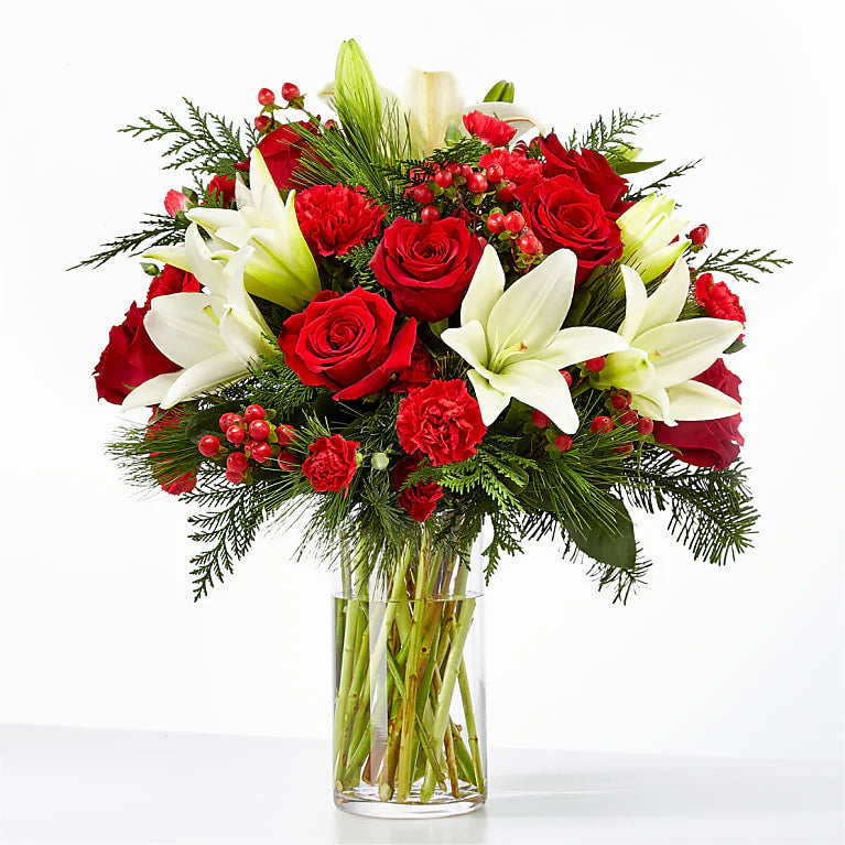 Festive red and white holiday bouquet featuring red roses, red carnations, white lilies, and hypericum berries, accented with lush evergreen sprigs. Arranged in a clear glass vase, this elegant floral display is perfect for Christmas decor or gifting