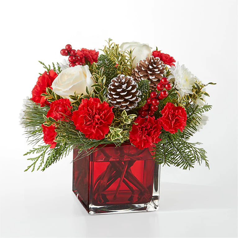 Festive red and white Christmas floral arrangement featuring red carnations, white roses, chrysanthemums, pinecones, hypericum berries, and fresh greenery, displayed in a red glass cube vase. Perfect for holiday decor or gifting."