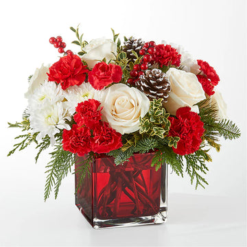 Festive red and white Christmas floral arrangement featuring red carnations, white roses, chrysanthemums, pinecones, hypericum berries, and fresh greenery, displayed in a red glass cube vase. Perfect for holiday decor or gifting."
