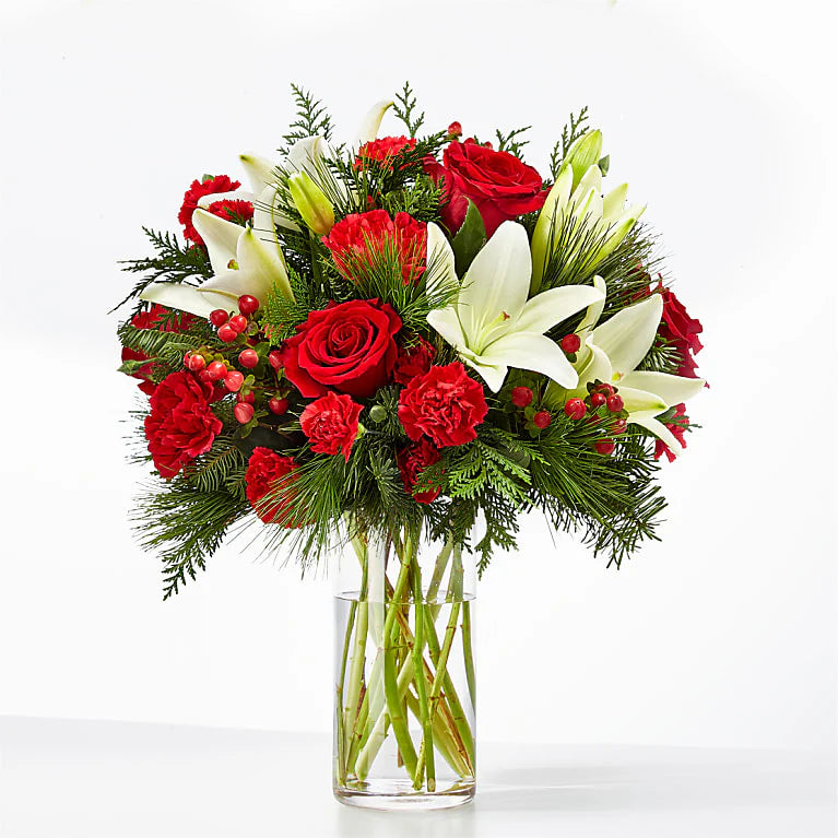 Festive red and white holiday bouquet featuring red roses, red carnations, white lilies, and hypericum berries, accented with lush evergreen sprigs. Arranged in a clear glass vase, this elegant floral display is perfect for Christmas decor or gifting