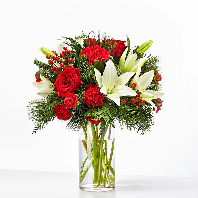 Festive red and white holiday bouquet featuring red roses, red carnations, white lilies, and hypericum berries, accented with lush evergreen sprigs. Arranged in a clear glass vase, this elegant floral display is perfect for Christmas decor or gifting