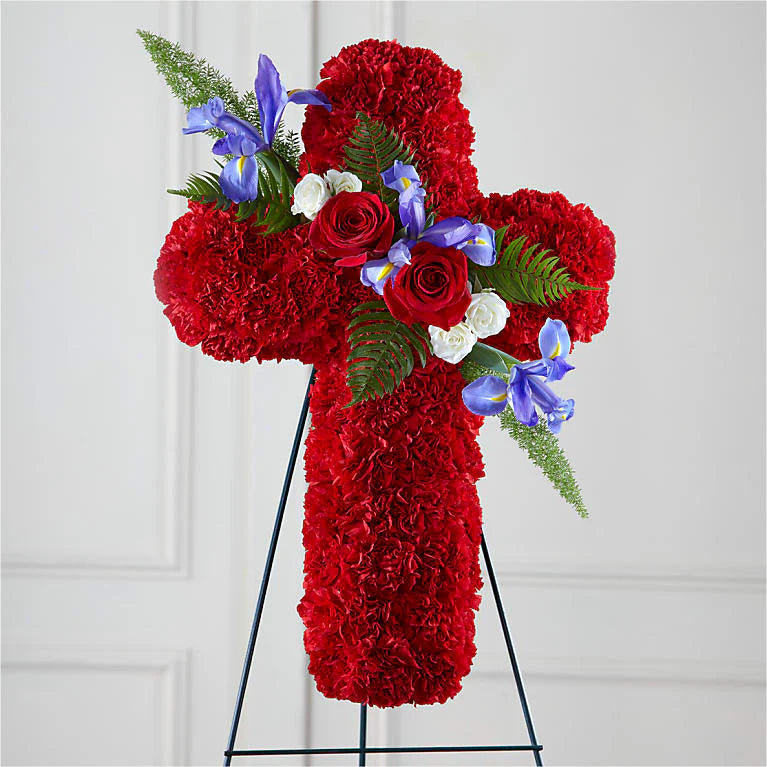 Cross-shaped floral arrangement made with red carnations, accented by red roses, white spray roses, blue irises, and lush greenery, displayed on a black stand. A vibrant and meaningful tribute symbolizing love, faith, and remembrance for a funeral or memorial service