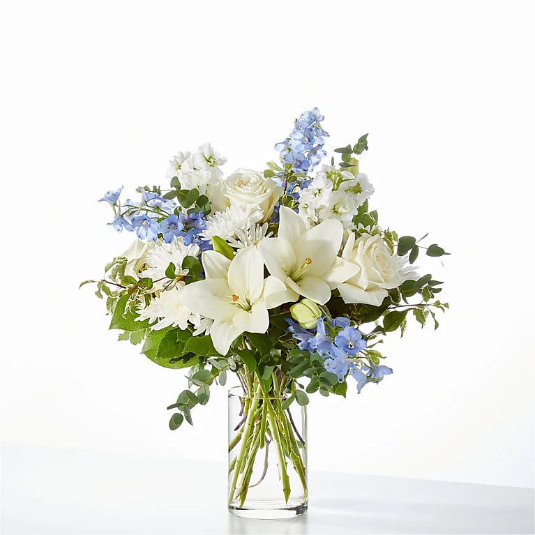 Elegant blue and white flower arrangement with white lilies, ivory roses, carnations, and blue delphiniums in a clear glass vase, perfect for sympathy, new baby gifts, or home decor