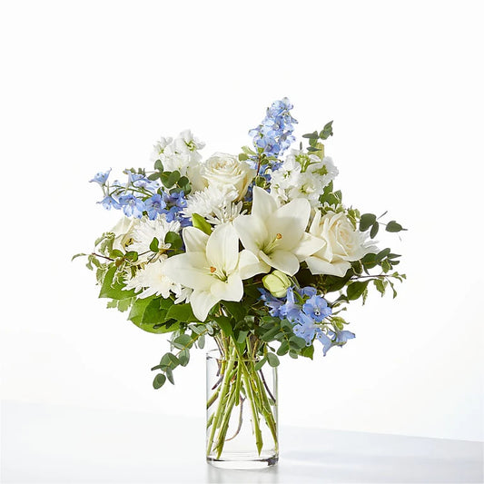 Elegant blue and white flower arrangement with white lilies, ivory roses, carnations, and blue delphiniums in a clear glass vase, perfect for sympathy, new baby gifts, or home decor