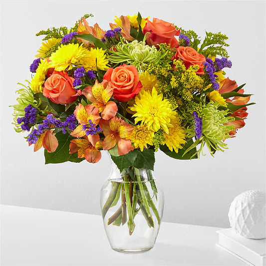 Bright mixed bouquet featuring vibrant orange roses, yellow chrysanthemums, peach alstroemeria, green spider mums, and purple statice, arranged in a clear glass vase with fresh green foliage. The colorful floral display exudes a cheerful and festive feel, perfect for any celebration 