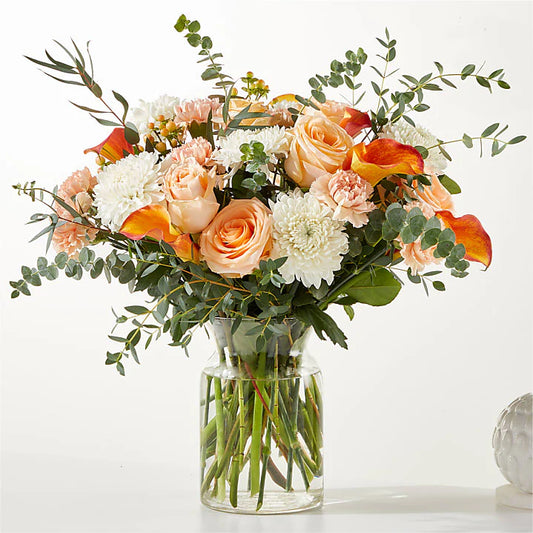 A close-up of a bouquet with soft peach-colored roses, vibrant gerbera daisies, and elegant lilies, surrounded by fresh greenery. The arrangement features warm, inviting hues of peach, pink, and white, creating a cheerful and delicate floral display.