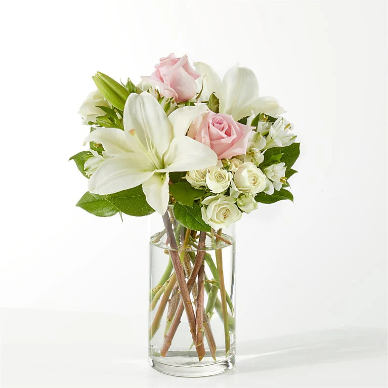 Elegant floral arrangement featuring white lilies, soft pink roses, and light daisies in a clear vase, symbolizing peace and remembrance