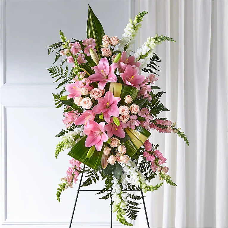 Elegant Embrace Standing Spray featuring a stunning arrangement of white lilies, snapdragons, and lush greenery, designed in a cascading style for a funeral or memorial service. The white blooms symbolize peace and eternal love, offering a serene and comforting tribute to honor a loved one.
