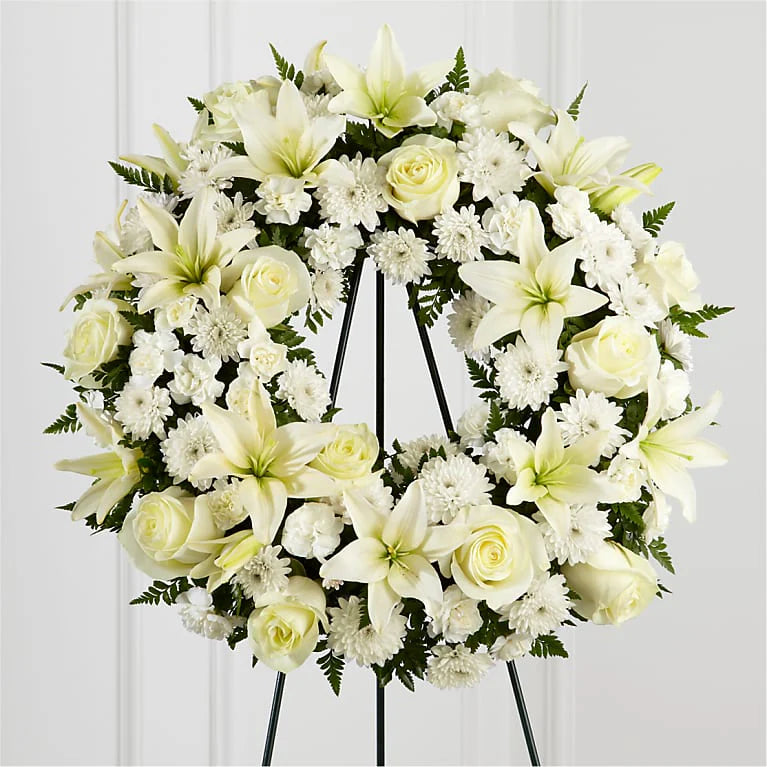 Round white floral wreath featuring white lilies, roses, and chrysanthemums, arranged with lush green ferns. The design is elegant and serene, symbolizing peace, love, and remembrance. The wreath is displayed on a black metal stand against a light background 