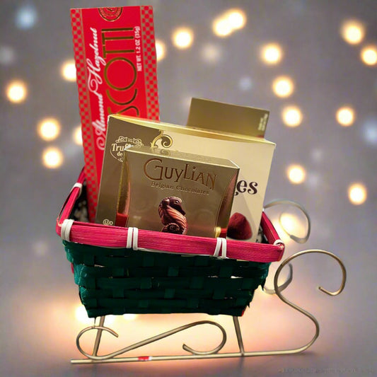 A festive holiday gift basket arranged in a sleigh-shaped woven container with green and red accents. The basket includes Highland Almond Biscotti, Guylian Belgian chocolates, and other gourmet treats. Perfect for Christmas, seasonal celebrations, or corporate gifting, this elegant arrangement spreads festive cheer and holiday warmth. 