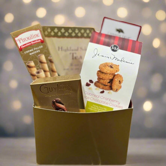 A gold gift box containing an assortment of snacks and treats, including Pirouline chocolate hazelnut crème-filled wafers, a box of Highland Specialty Tea, Guylian Belgian chocolates, and J&M oatmeal cranberry white chocolate cookies. The arrangement is neatly organized, making it a perfect gift for tea and snack lovers.