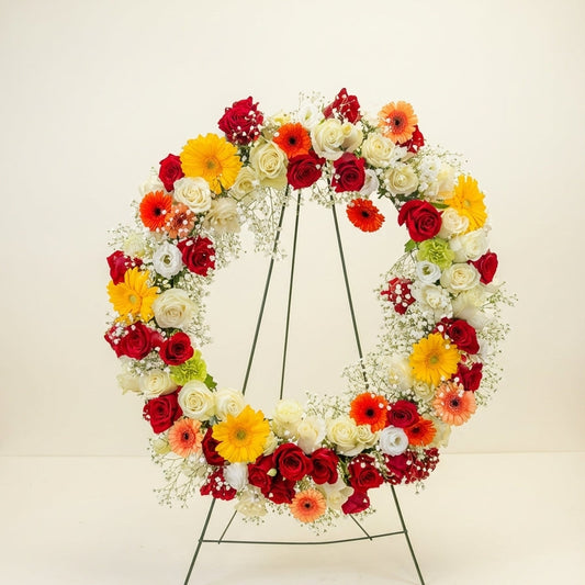 Floral wreath with red, yellow, and white flowers on a stand against a light background