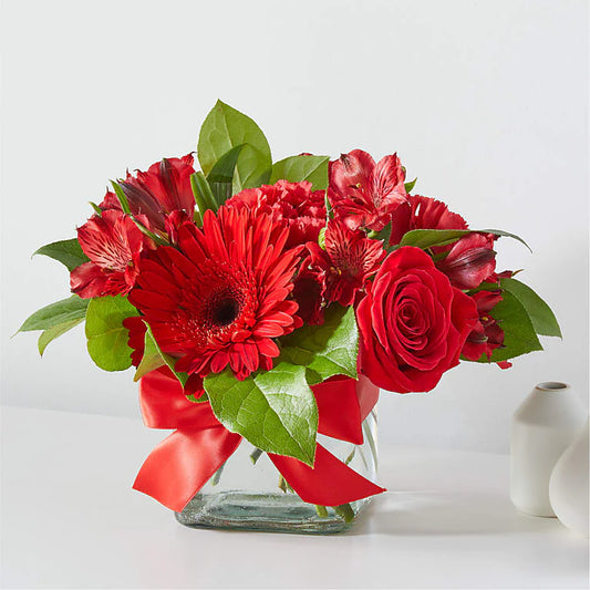 Vibrant red flower arrangement featuring bold red roses, carnations, and lush greenery, beautifully arranged in a decorative vase.