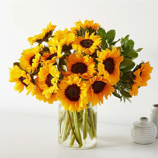 Bright sunflower bouquet arranged in a clear glass vase, featuring vibrant yellow blooms and green foliage.

