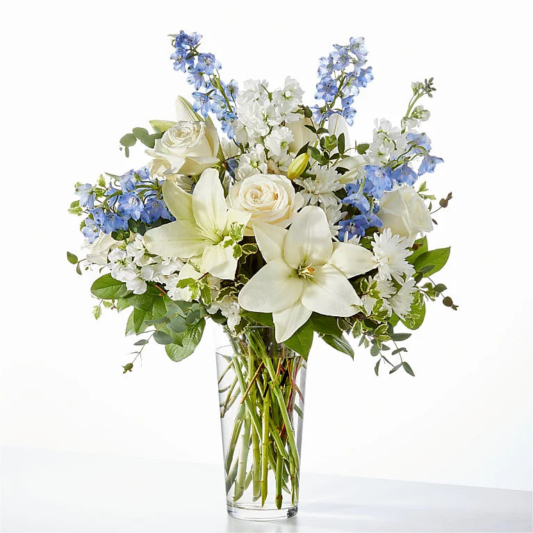 Elegant blue and white flower arrangement with white lilies, ivory roses, carnations, and blue delphiniums in a clear glass vase, perfect for sympathy, new baby gifts, or home decor
