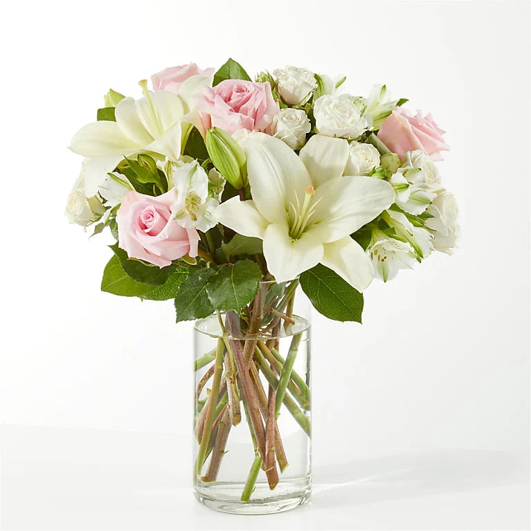 Elegant floral arrangement featuring white lilies, soft pink roses, and light daisies in a clear vase, symbolizing peace and remembrance